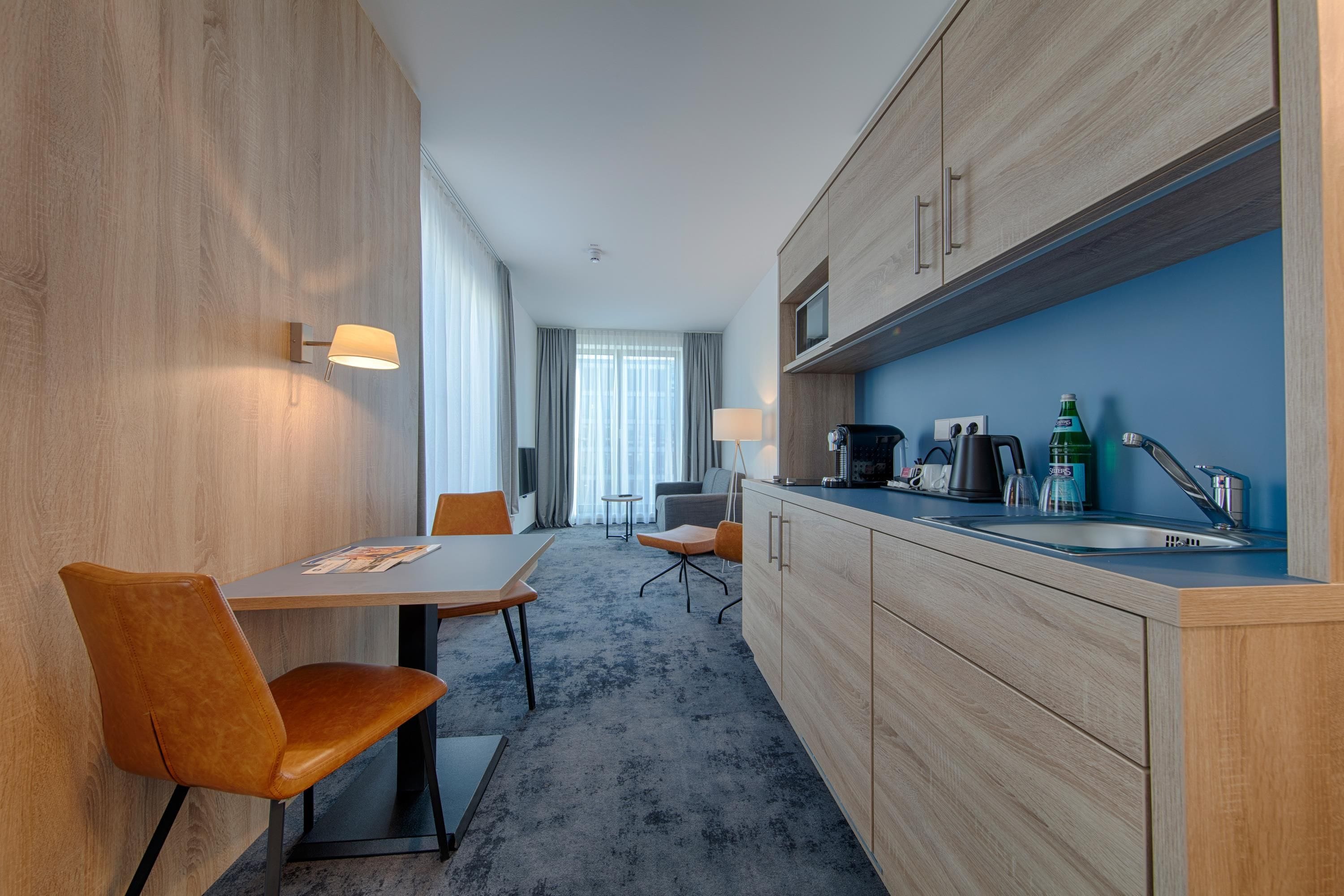 A modern hotel room with a dining area, kitchenette, and a view of the outdoors.