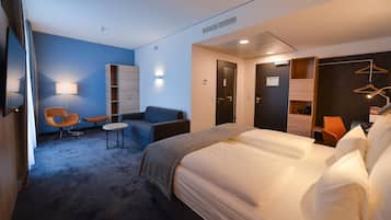A hotel room with a bed, a seating area with a chair and a small table, a wardrobe, and a desk with a lamp.
