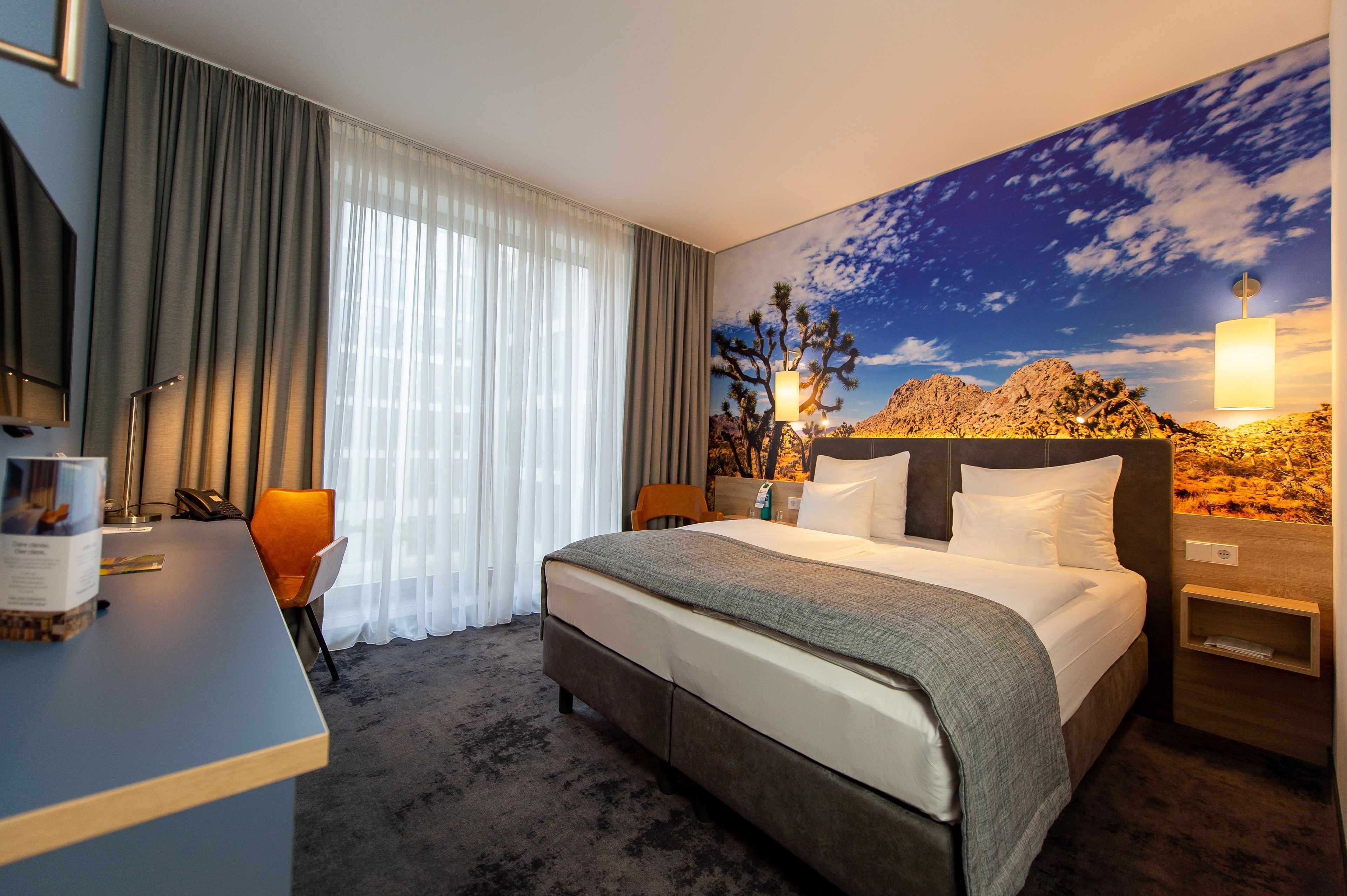 Ein Hotelzimmer mit einem Bett, einem Schreibtisch, einem Sessel, einem Nachttisch und einem großen Fenster mit Vorhängen.