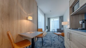 A modern hotel room with a wooden accent wall, a dining area with a table and chairs, a kitchenette, and large windows with curtains. - Best Western Hotel Wiesbaden (Wiesbaden)