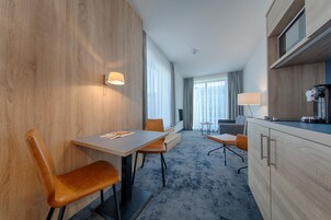 Habitación de hotel moderna con una pared de acento de madera, zona de comedor con mesa y sillas, kitchenette y ventanas amplias con cortinas.