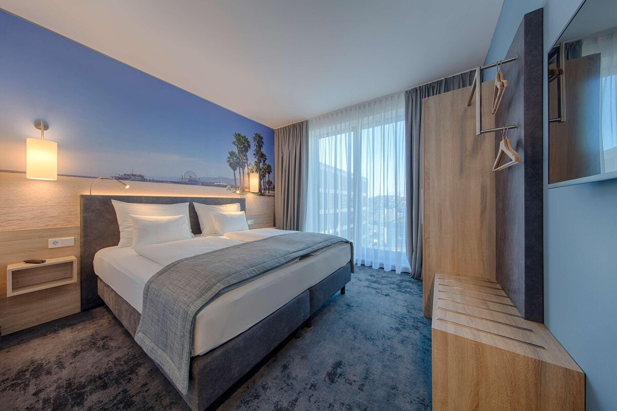 Habitación de hotel moderna con una cama grande, una mesita de noche, un banco de madera, un televisor y vistas al mar.