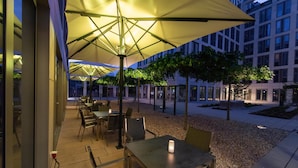 Un área de comedor al aire libre con mesas y sombrillas, rodeada de edificios y árboles.
