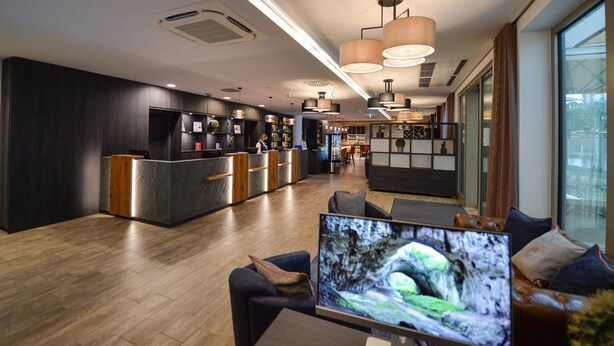 Un moderno vestíbulo de hotel con una recepción, zona de estar y una gran pantalla de televisión que muestra una escena natural.