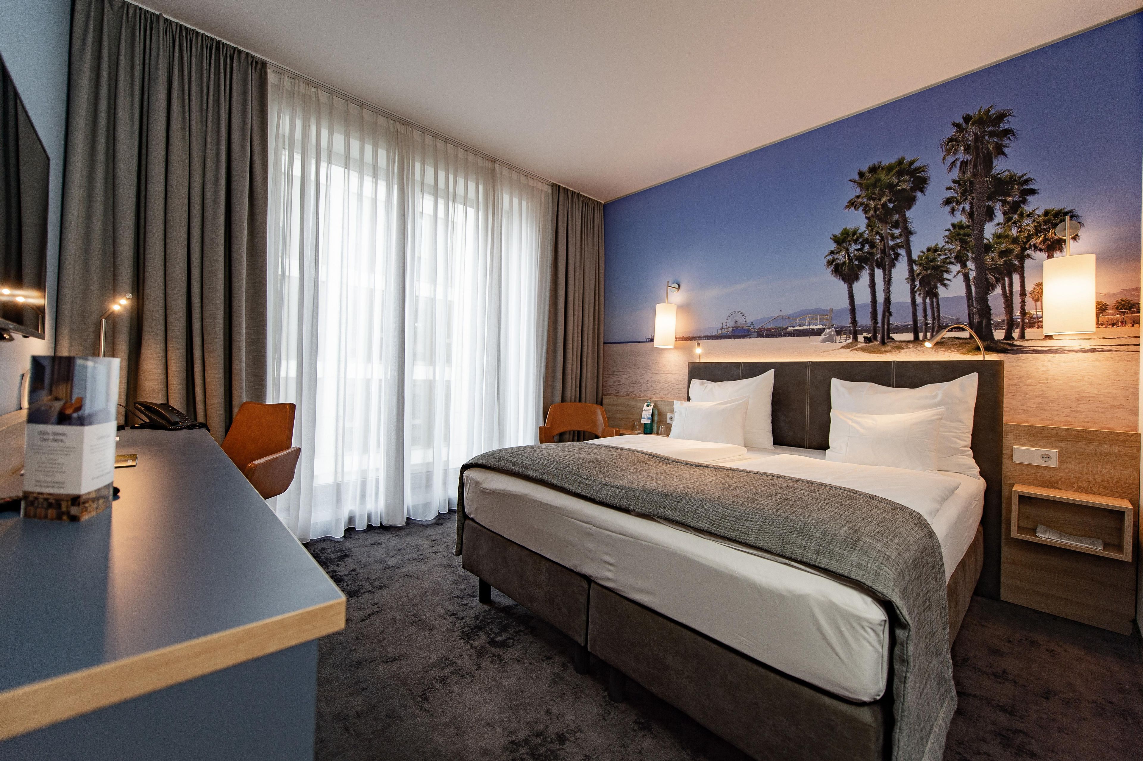 A hotel room with a bed, a desk, a chair, a nightstand, and a wall with a beach view.
