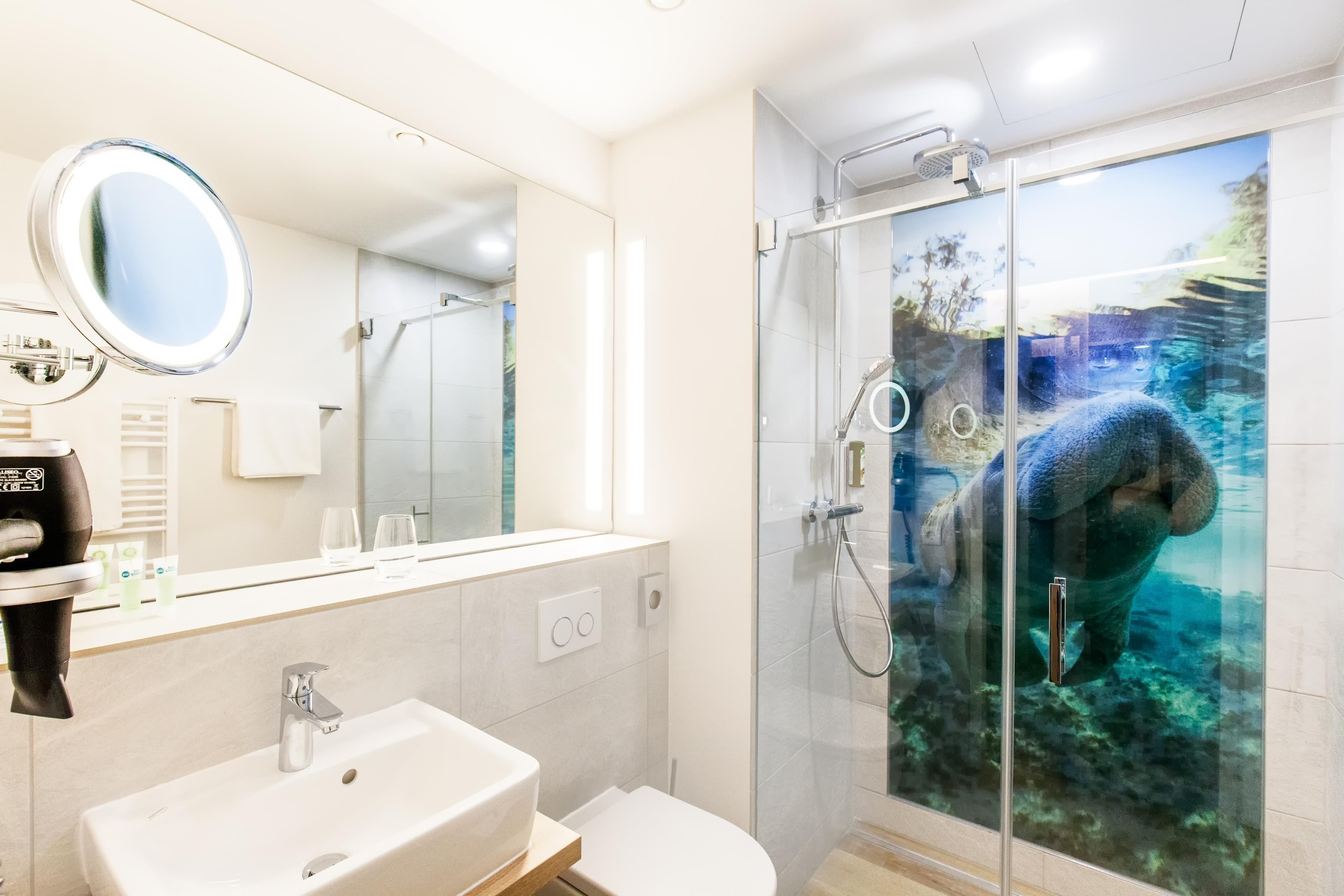 Un baño moderno con un espejo grande, un lavabo, un inodoro y una ducha con mampara que presenta un mural de una criatura marina.