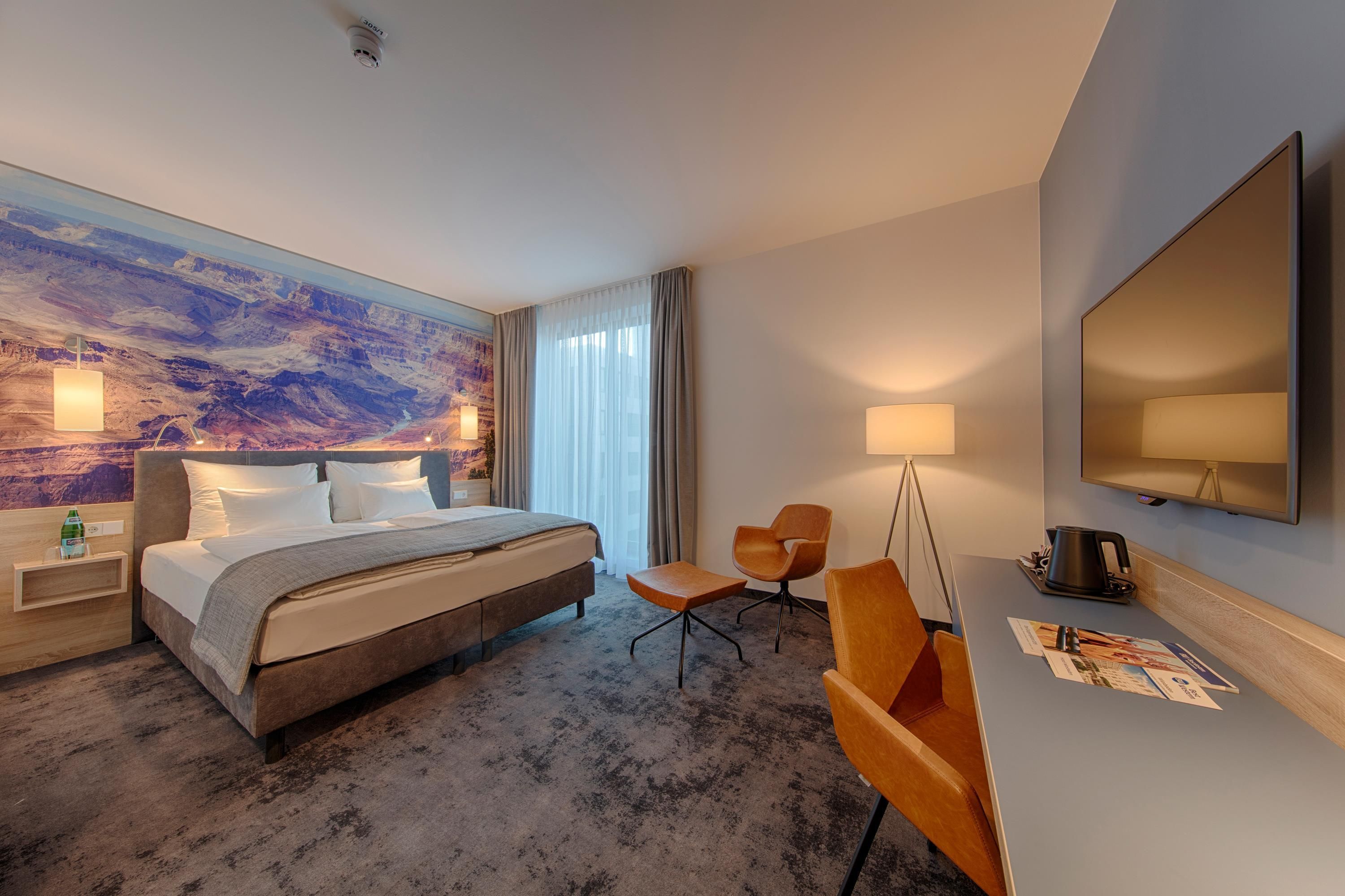Ein Hotelzimmer mit Bett, Schreibtisch, Stuhl, Flachbildfernseher und einem großen Fenster mit Vorhängen.
