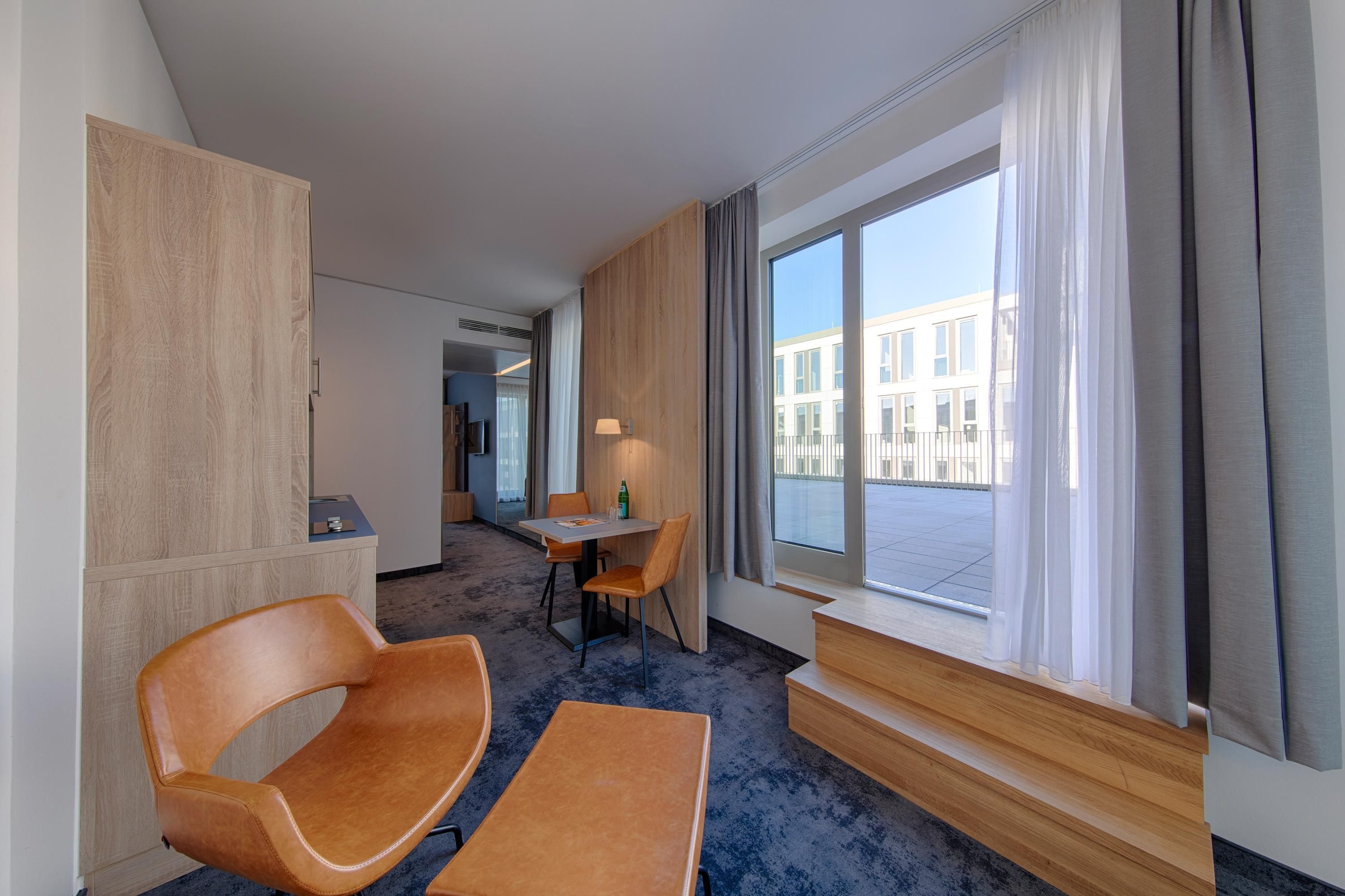 A hotel room with a seating area, a table, a window with curtains, and a view of the outside building.