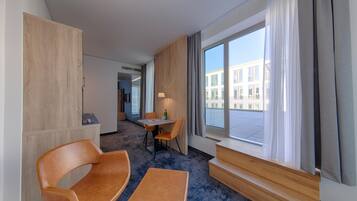A hotel room with a seating area, a table, a window with curtains, and a view of the outside building.