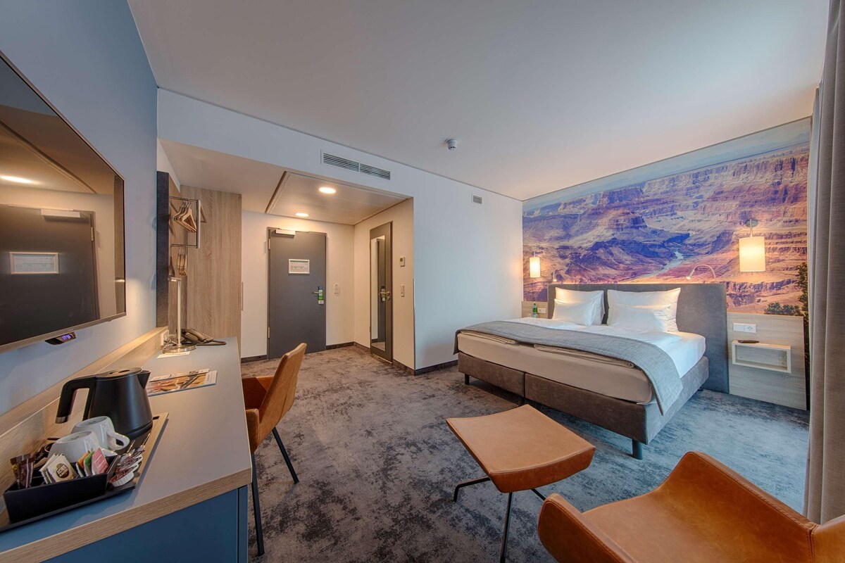 Una habitación de hotel con una cama, un escritorio con lámpara, una silla, una mesita y un mural grande en la pared.