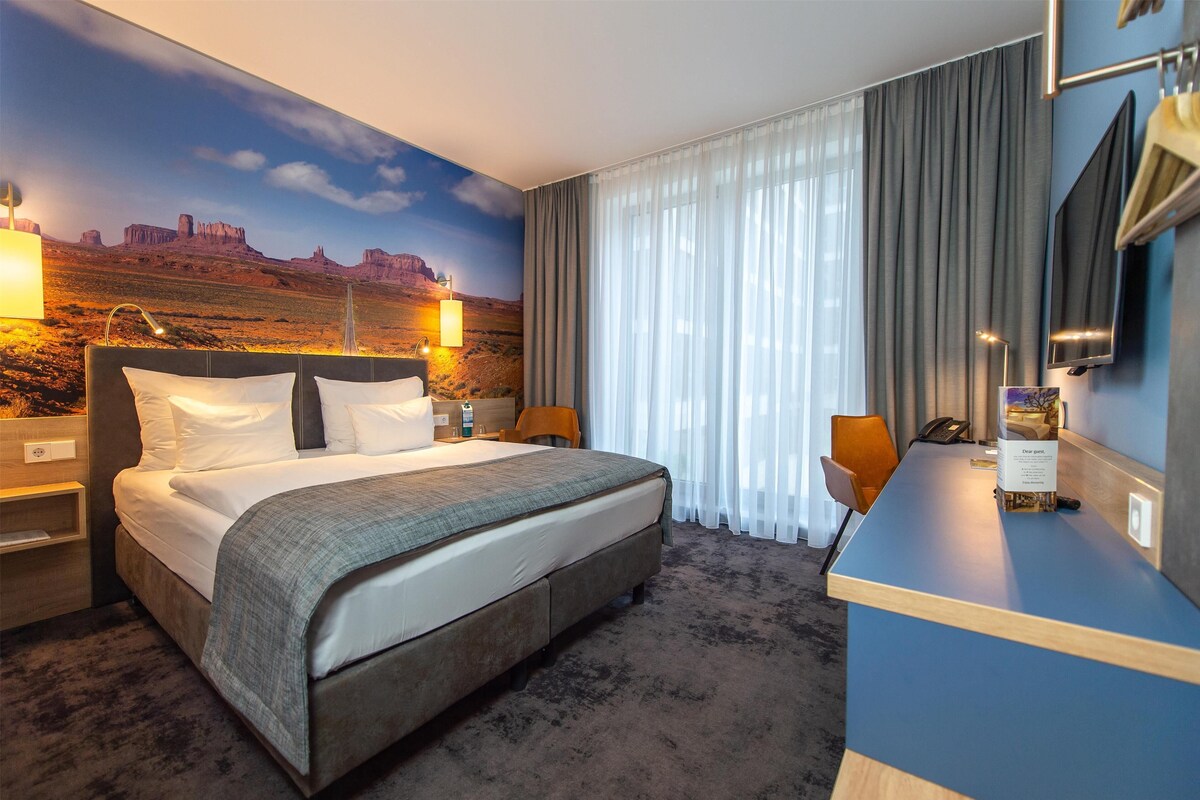 Habitación de hotel con una cama grande, un escritorio, una silla, un televisor y un mural de un paisaje desértico.