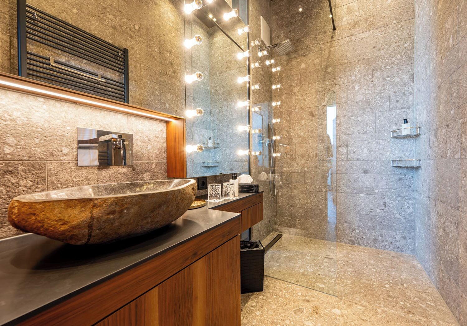 Un baño moderno con un lavamanos de piedra grande, tocador de madera y zona de ducha con mampara de vidrio.