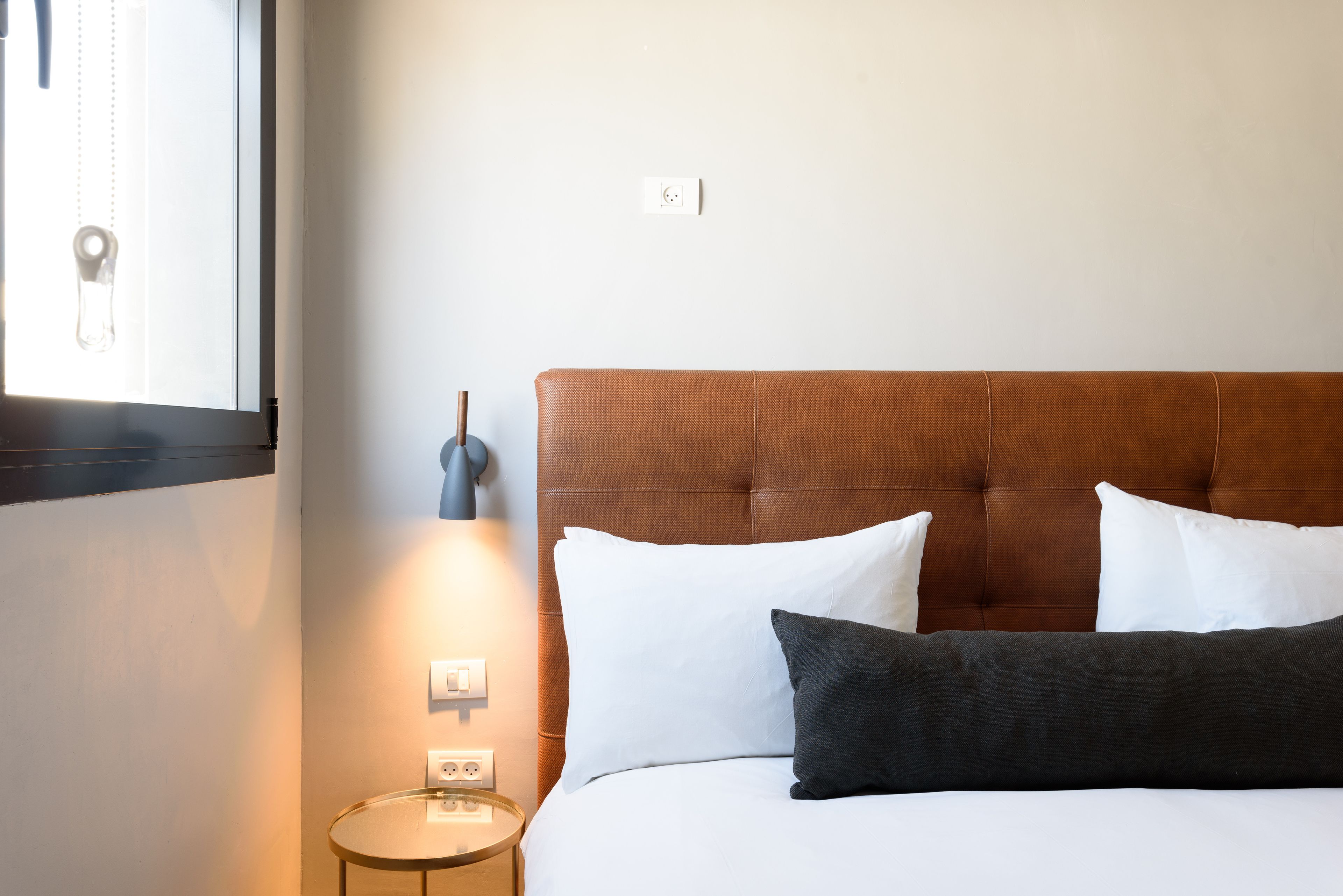 A neatly made bed with white and dark pillows, a round bedside table with a lamp, and a window with blinds.