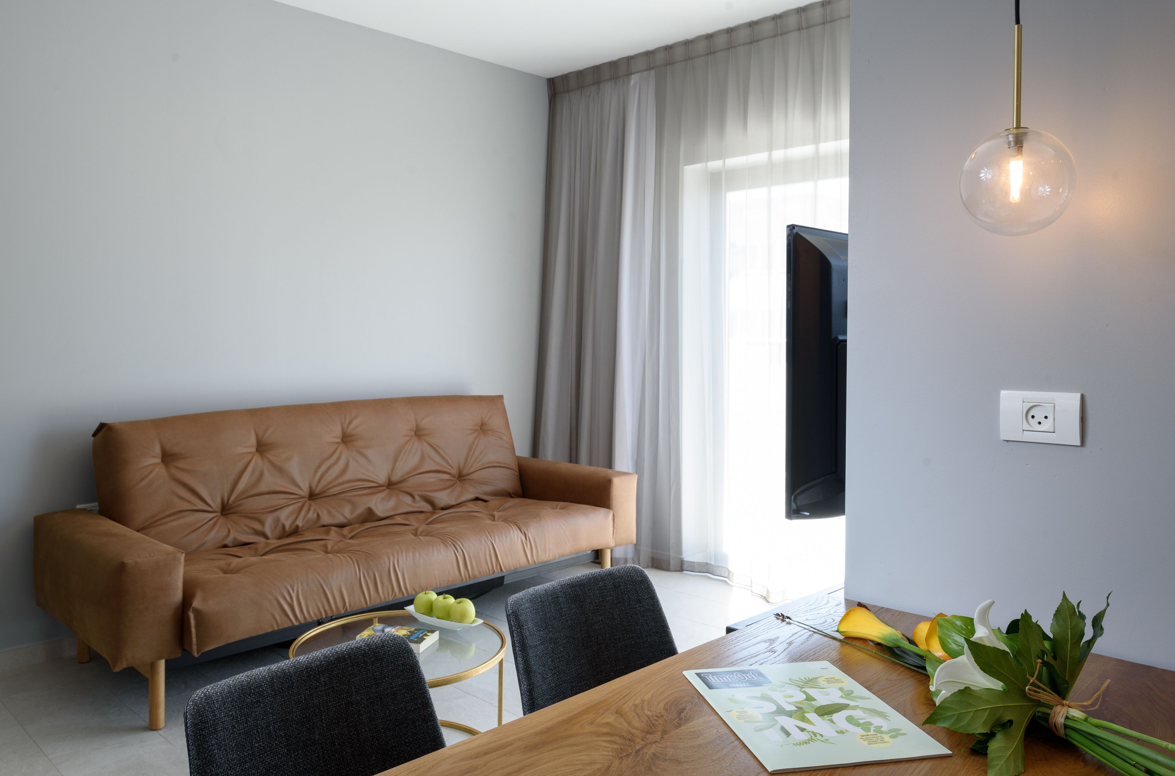 A modern living room with a brown leather sofa, a wooden dining table with chairs, a glass pendant light, and a window with curtains.