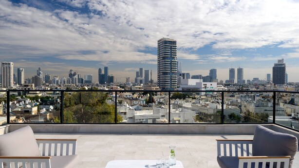 Une terrasse sur le toit offrant une vue sur la ville, avec une table agrémentée de verres et d’une bouteille, ainsi que deux chaises blanches dotées de coussins.