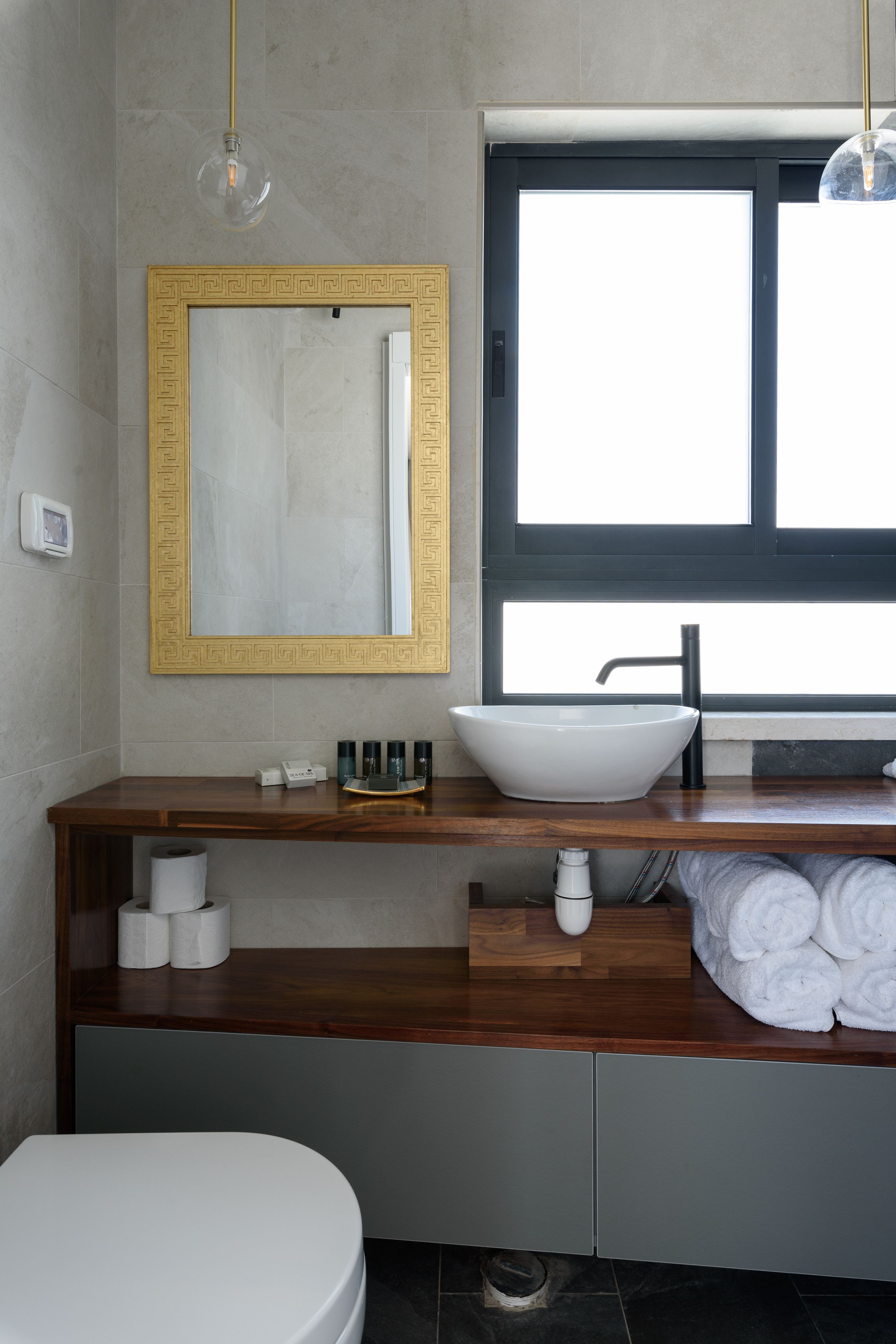 Un baño moderno con tocador de madera, un lavamanos blanco, un espejo grande con marco decorativo y una ventana que proporciona luz natural.