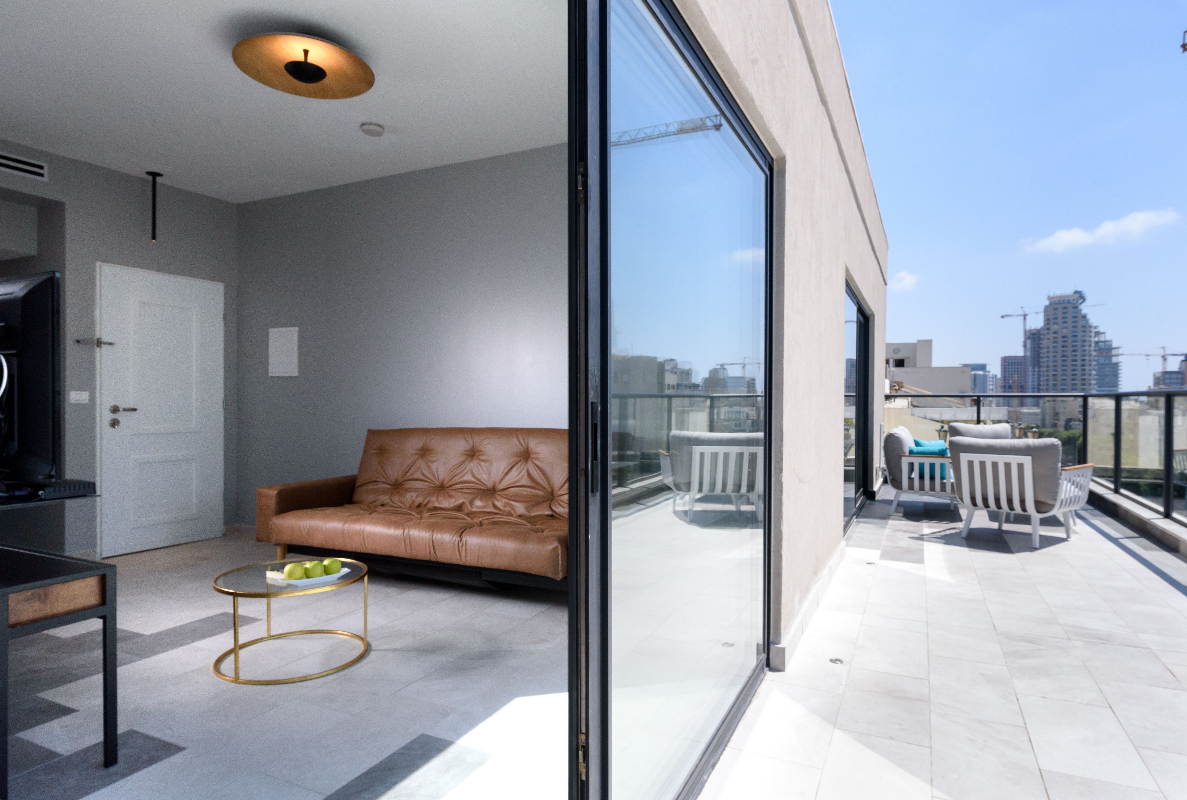 A modern living room with a leather sofa, a glass door leading to a balcony, and a view of the city skyline.
