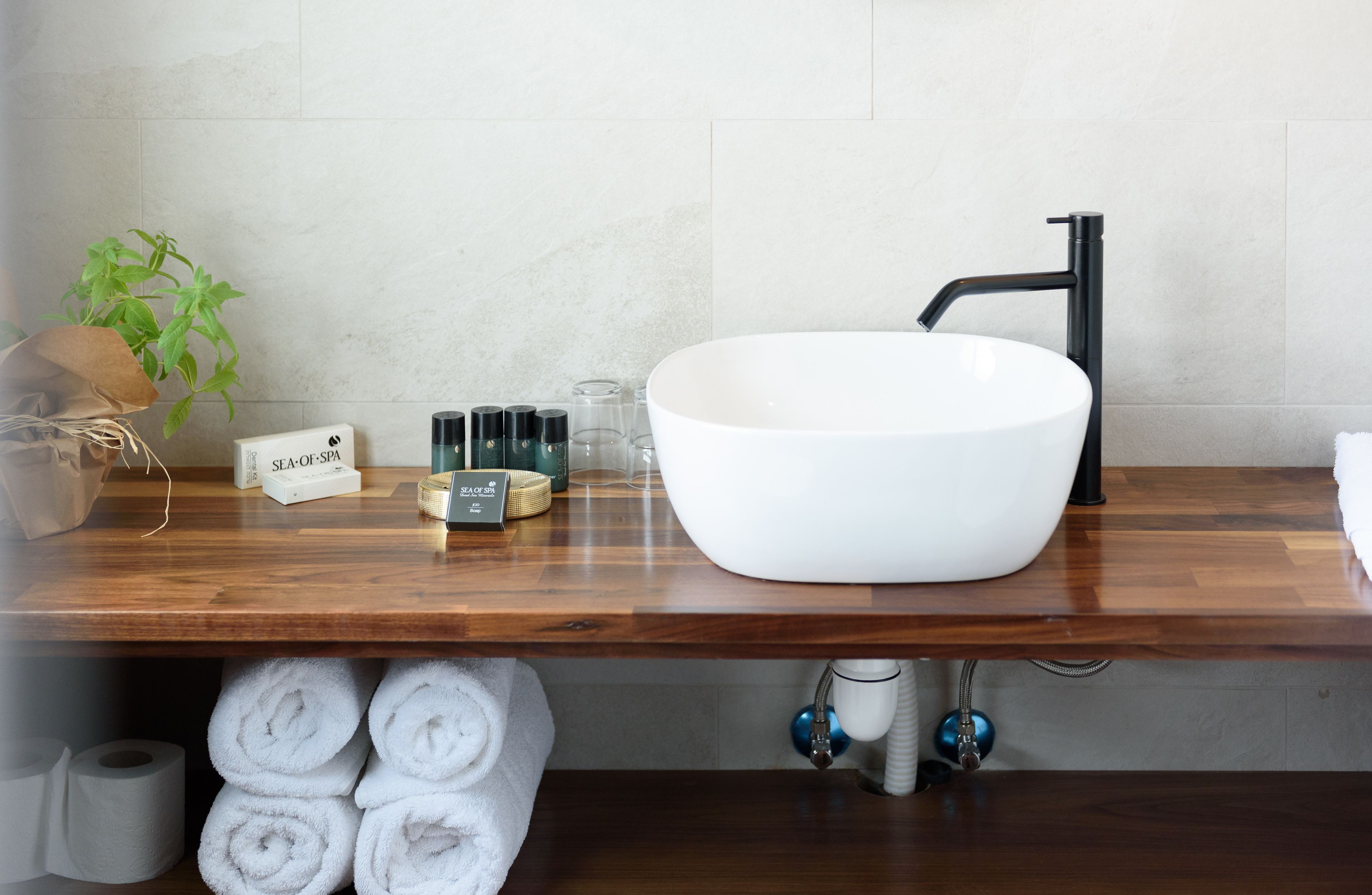 Un baño con un lavamanos grande de color blanco, grifo negro y encimera de madera. Hay toallas, una planta y artículos de aseo en la encimera.