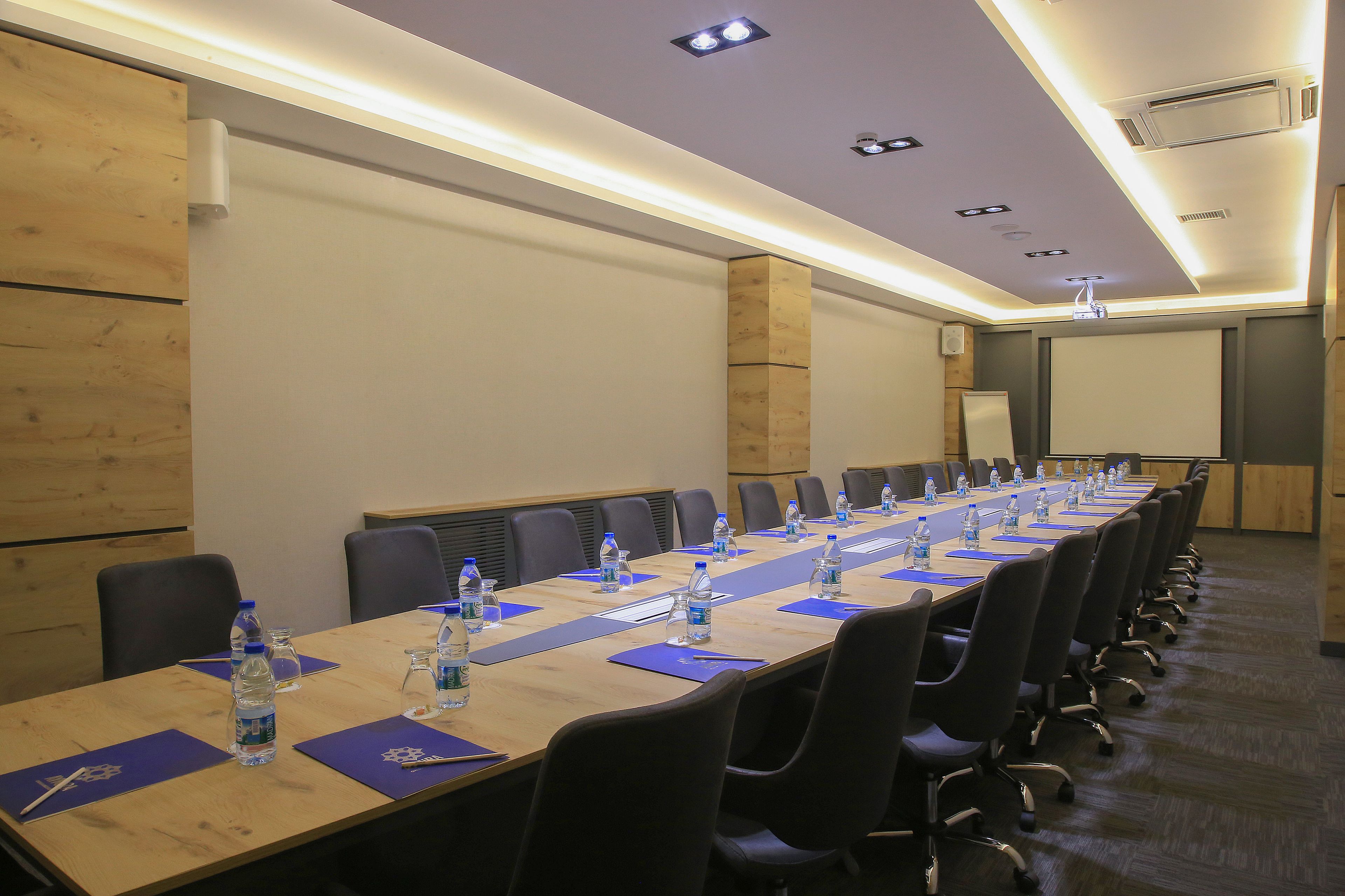 A conference room with a long table, chairs, water bottles, and a projector screen.