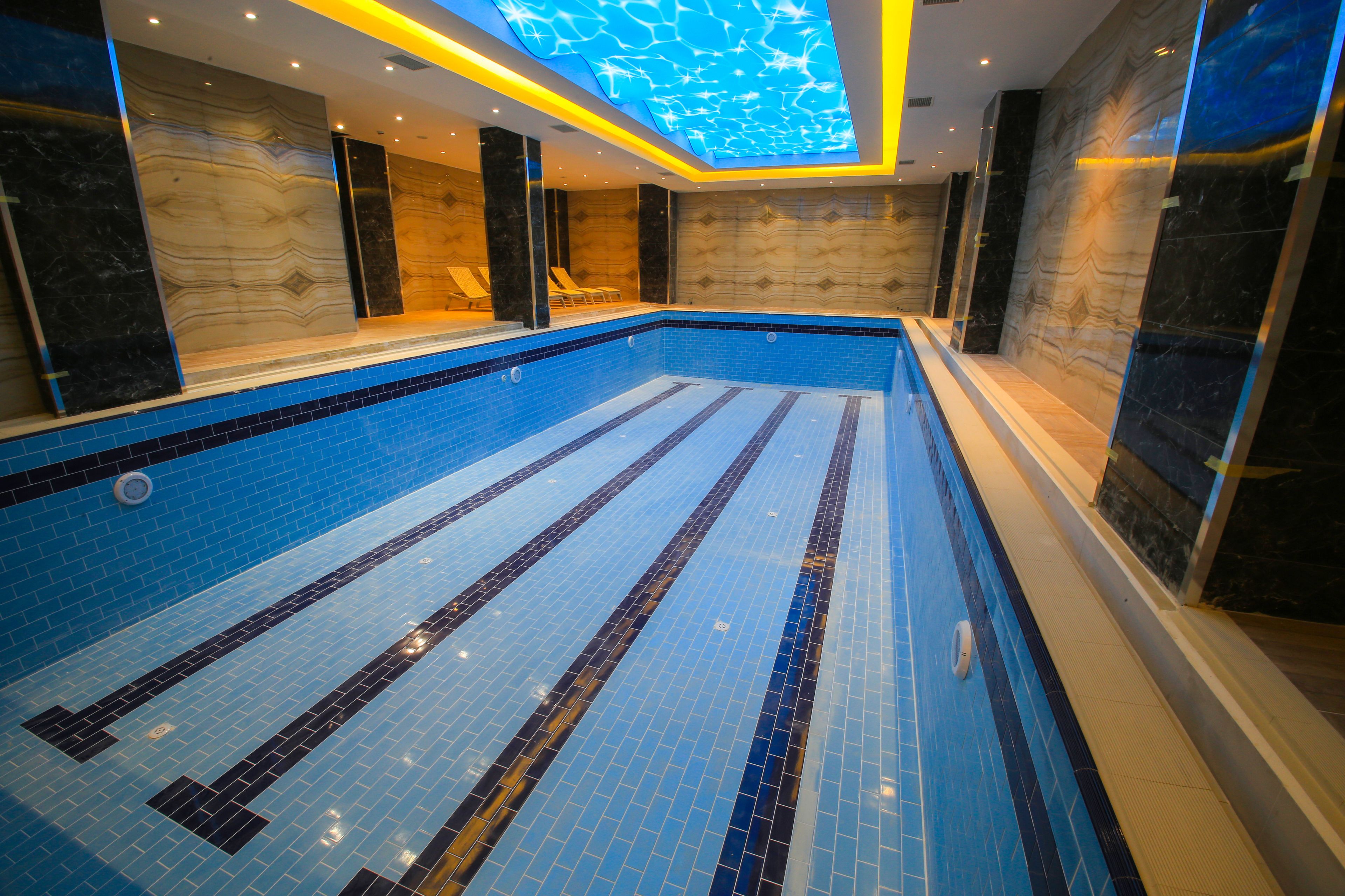 Una piscina con mosaicos azules y negros, una zona de estar hundida y un techo con un diseño luminoso.