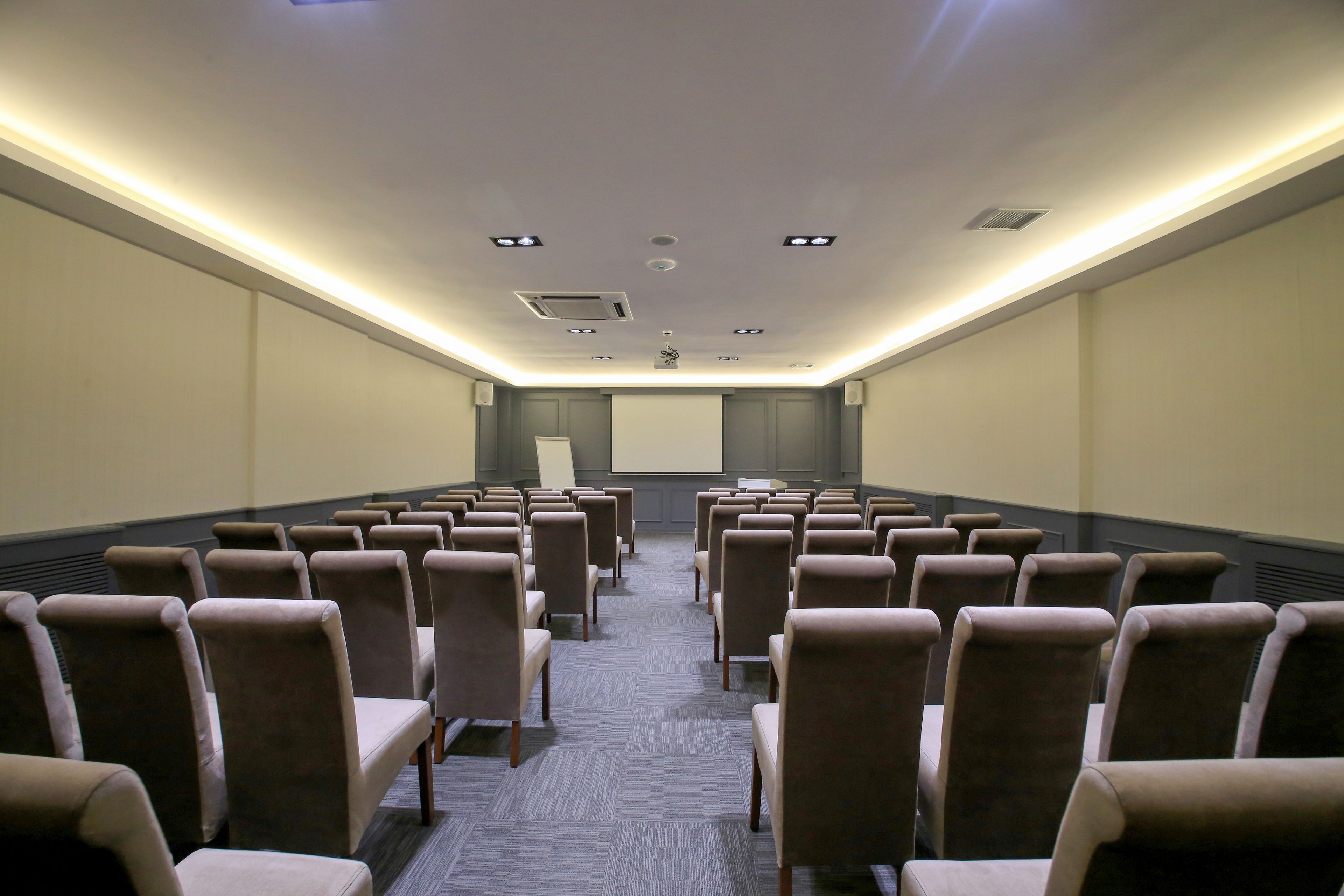 Uma sala de conferências com fileiras de cadeiras, uma tela de projeção e iluminação embutida no teto.