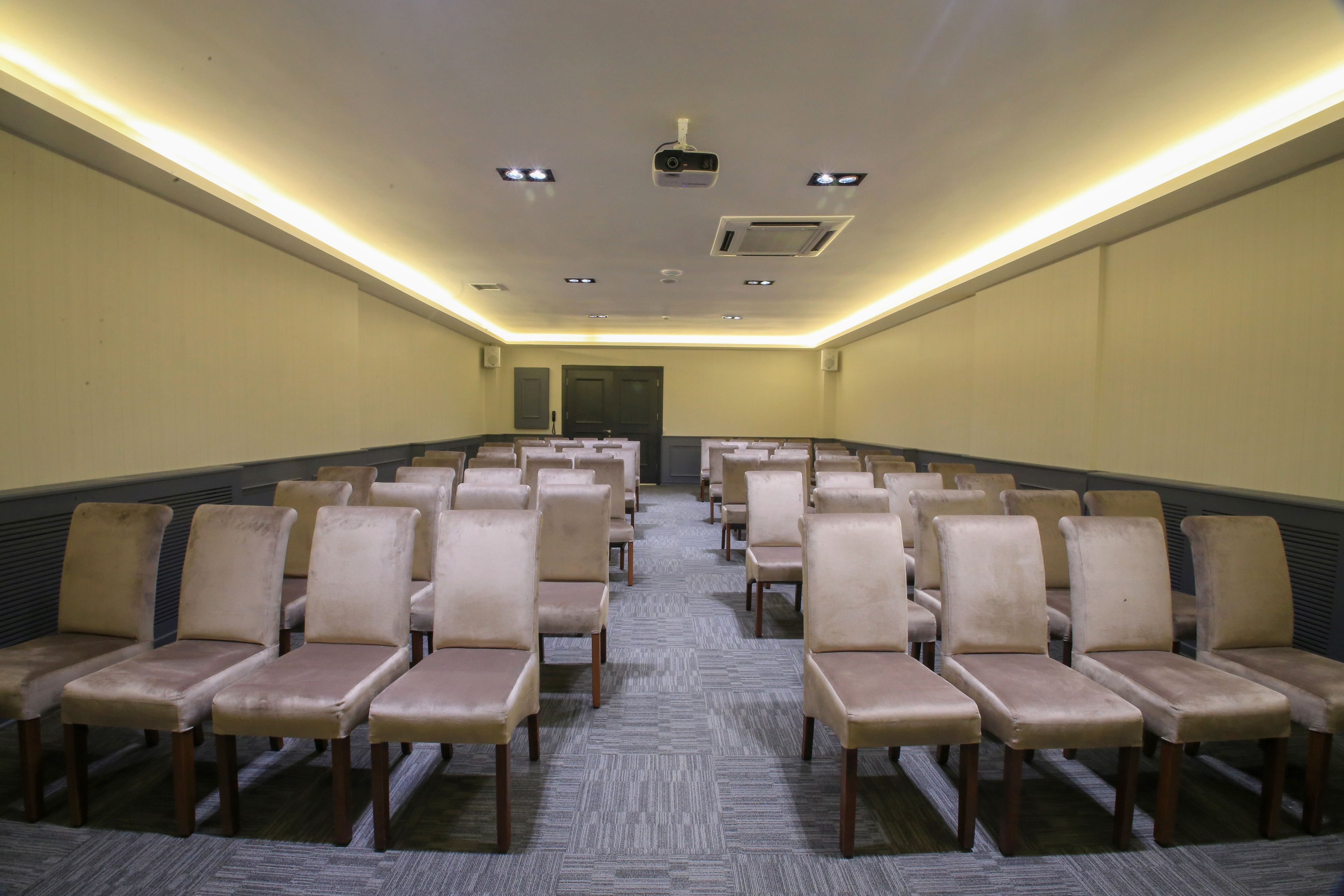 Un salón de conferencias con hileras de sillas color beige, suelo alfombrado, techo con iluminación empotrada y un proyector.