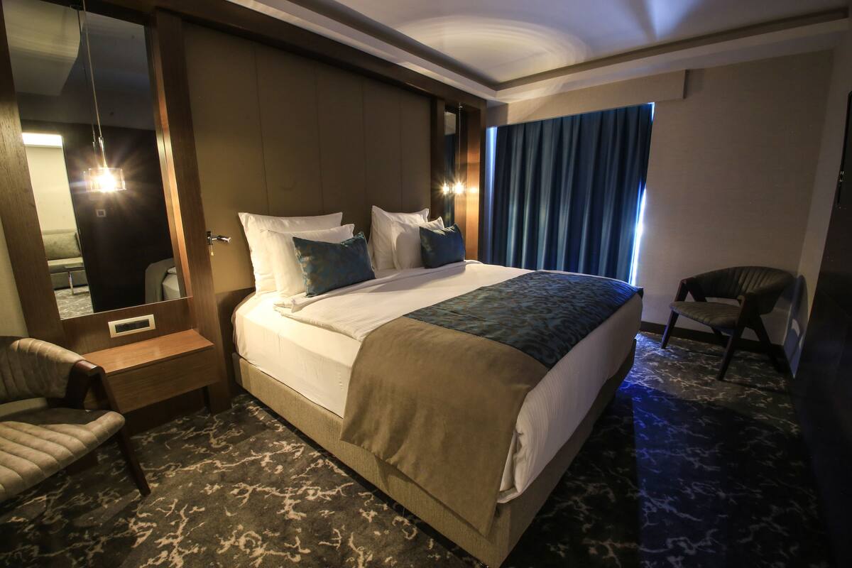 A hotel room with a large bed, a bedside table, a chair, a mirror, and a window with curtains.