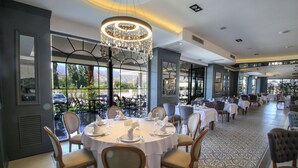 Un restaurant avec des tables rondes dressées pour le dîner, de grandes fenêtres donnant sur l’extérieur et un lustre suspendu au plafond.