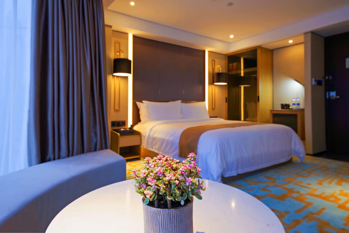 Een hotelkamer met een bed, nachtkastjes, een lamp, een kleintje tafeltje met een bloevas en een kamerbreed tapijt.