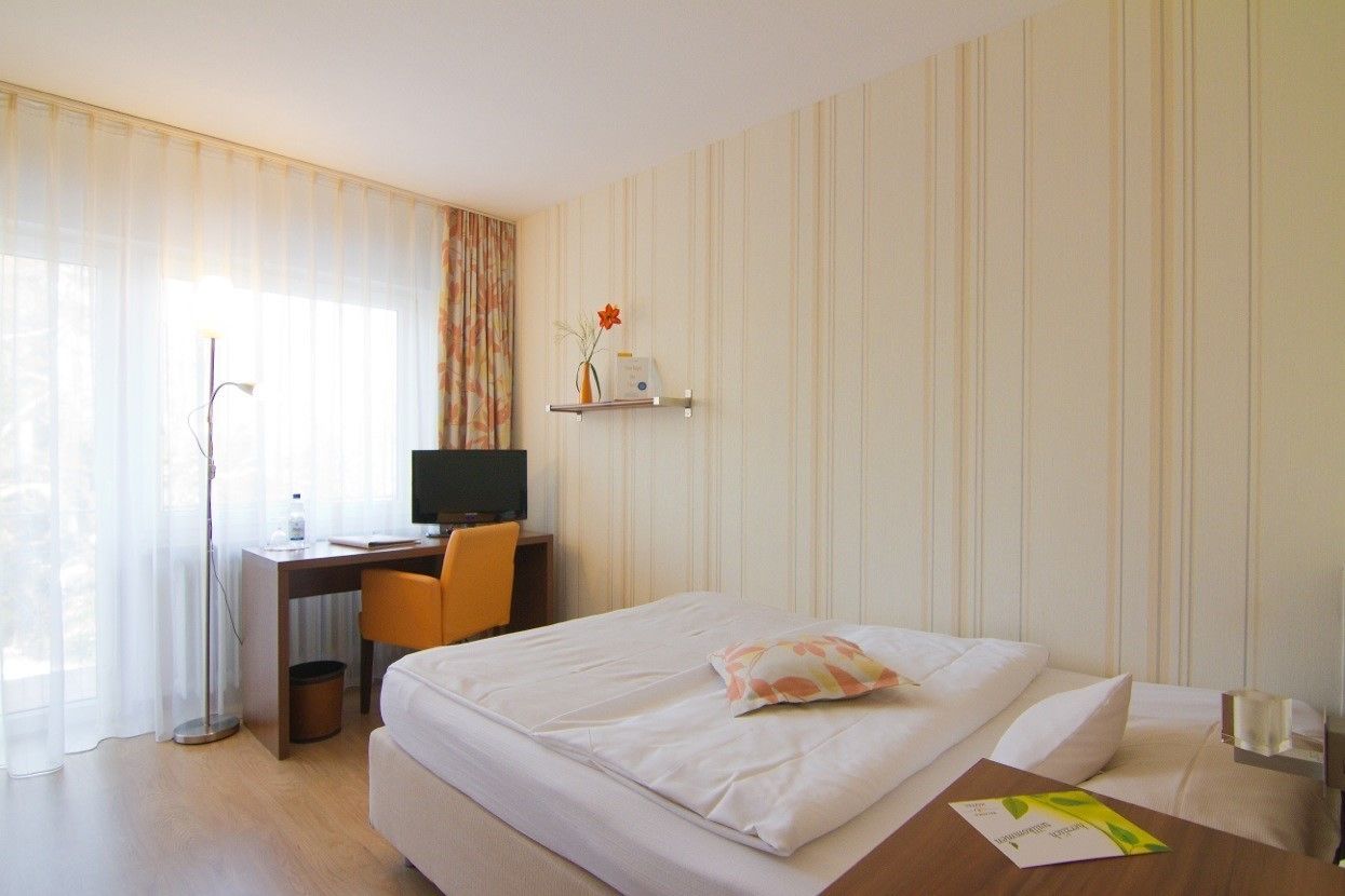 A hotel room with a bed, a desk with a computer, a chair, and a small shelf with a vase and a flower.