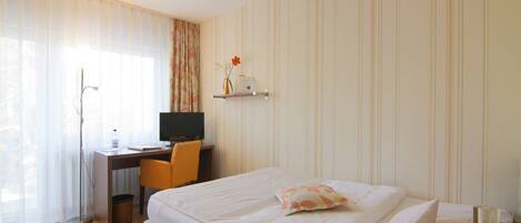 A hotel room with a bed, a desk with a computer, a chair, and a small shelf with a vase and a flower.