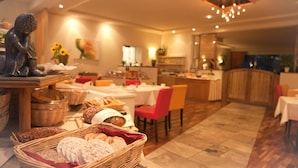 A modern dining area with a variety of breads and pastries on a buffet table, a statue on the left, and a cozy seating arrangement with a red chair and a wooden table. - Balance-Hotel am Blauenwald (Badenweiler)