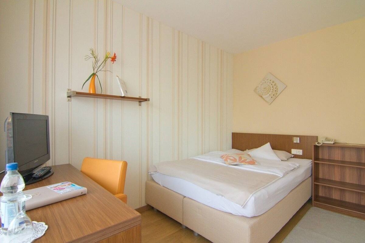 Een hotelkamer met een bed, een bureau met een computer, een stoel, een televisie en een plank met een bloevas.