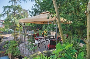 Ein überdachter Außenbereich mit Tischen und Stühlen, umgeben von üppiger Bepflanzung und einem Gartenweg.