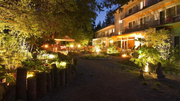 A well-lit outdoor area with a gravel path, surrounded by trees and plants, leading to a building with multiple balconies and windows. - Balance-Hotel am Blauenwald (Badenweiler)