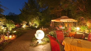An outdoor dining area with tables and chairs under a canopy, surrounded by lush greenery and illuminated by ambient lighting. - Balance-Hotel am Blauenwald (Badenweiler)