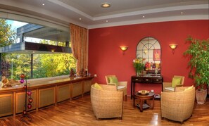 Ein Zimmer mit Holzboden, roten Wänden, einem großen Fenster mit Vorhängen, Korbmöbeln und einem kleinen Tisch mit Blumen.