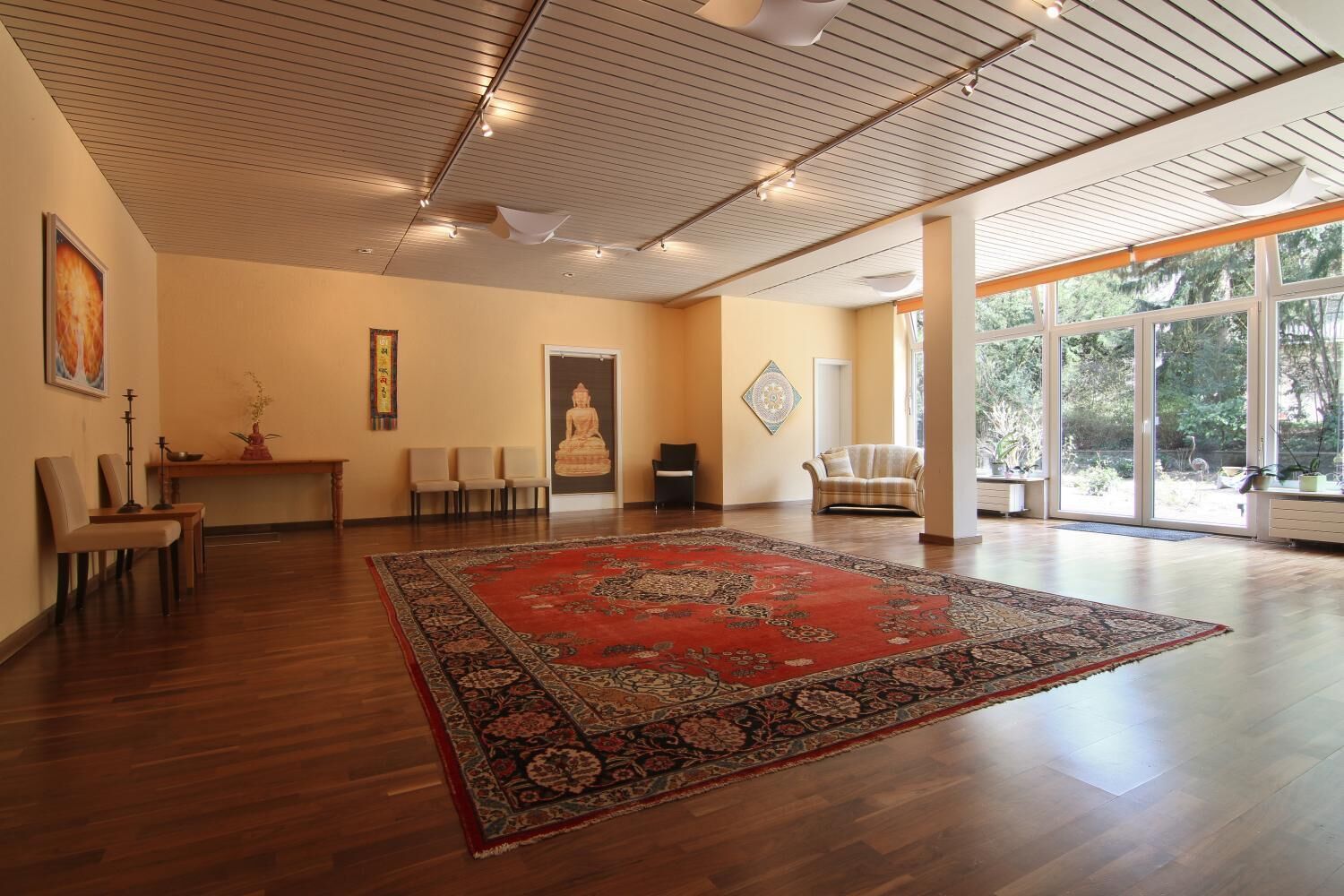 Une pièce spacieuse dotée d’un grand plancher en bois, d’un tapis rouge à motifs, d’une statue de Bouddha et offrant une vue sur la verdure grâce à de grandes fenêtres.