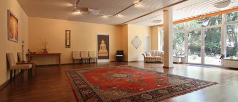 Une pièce spacieuse dotée d’un grand plancher en bois, d’un tapis rouge à motifs, d’une statue de Bouddha et offrant une vue sur la verdure grâce à de grandes fenêtres.