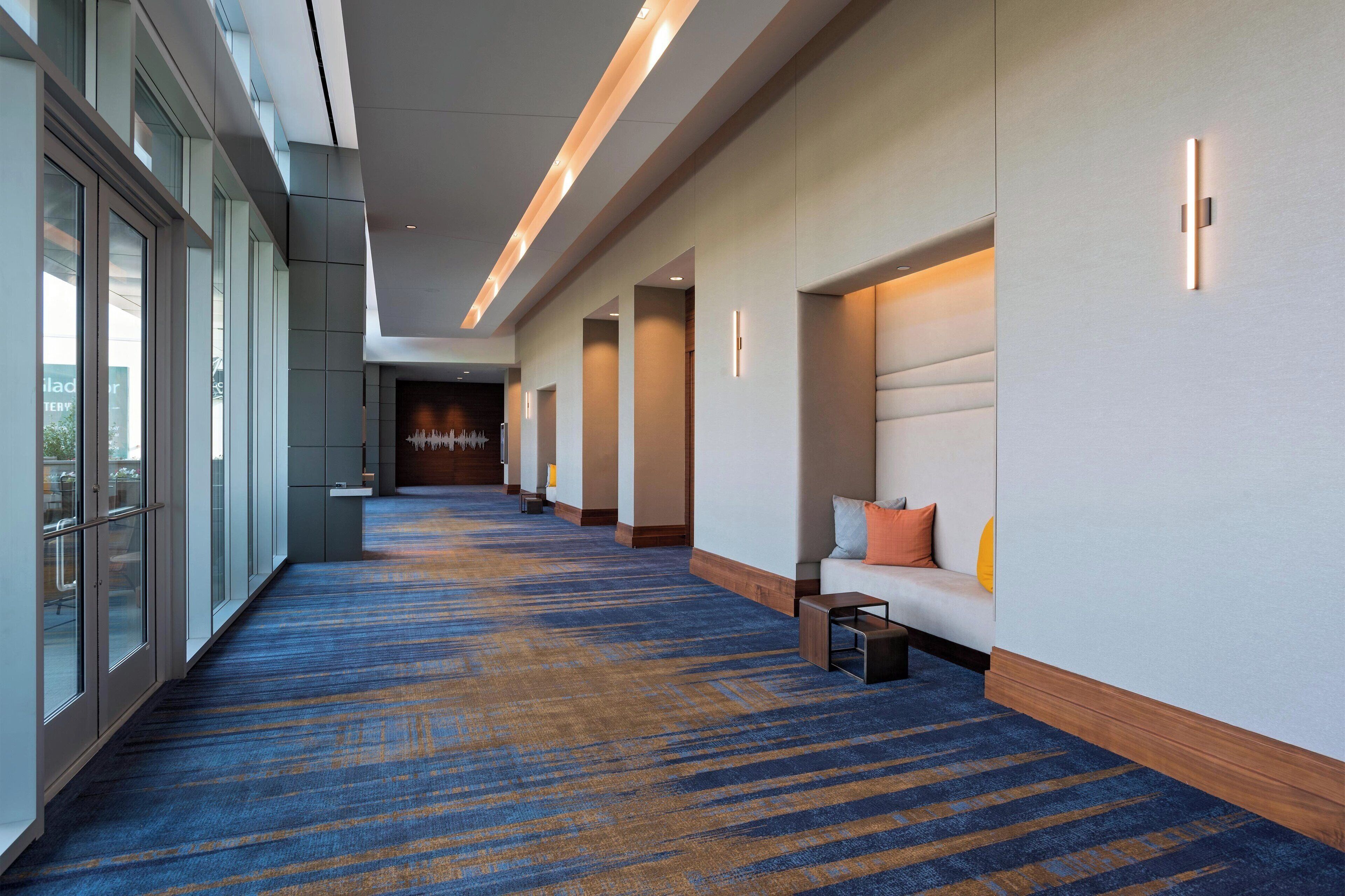 A hotel corridor with a blue carpet, seating area with cushions, and glass doors on one side.