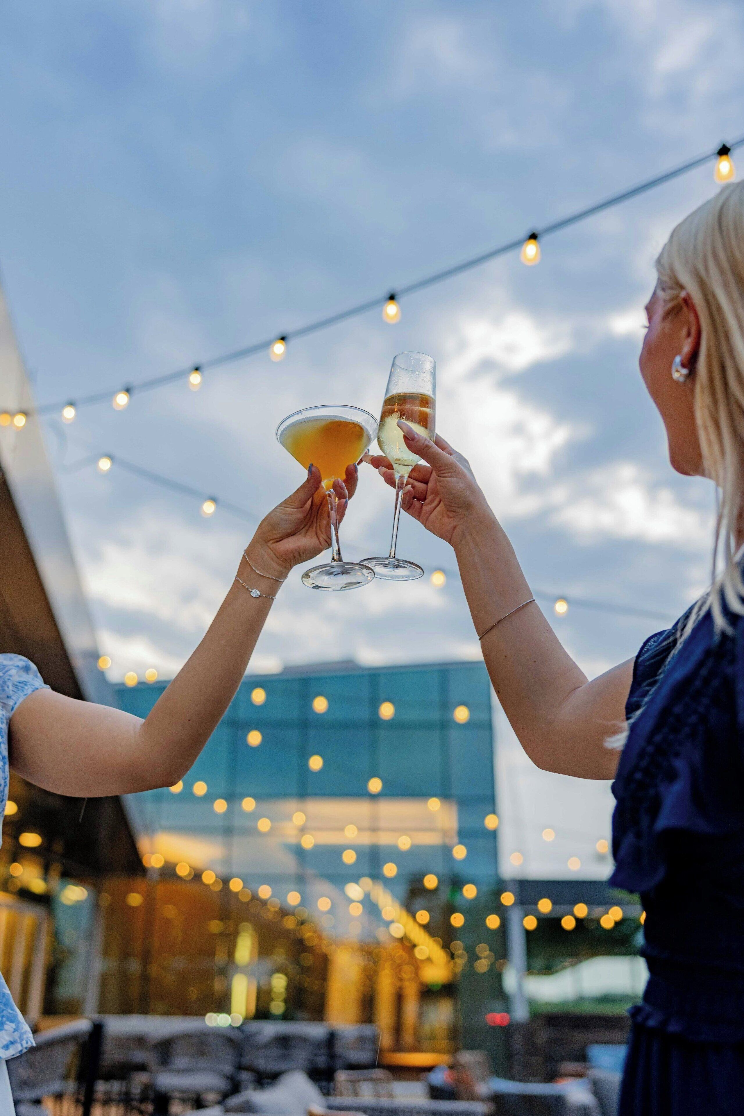 Two people toasting with drinks in an outdoor setting with string lights and a modern building in the background.