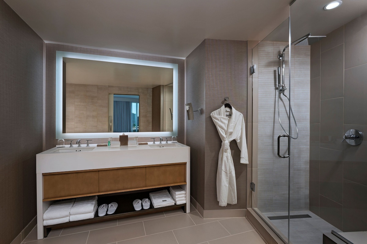 Un baño moderno con un espejo grande, lavamanos dobles, una bata colgada y una zona de ducha con cerramiento de vidrio.