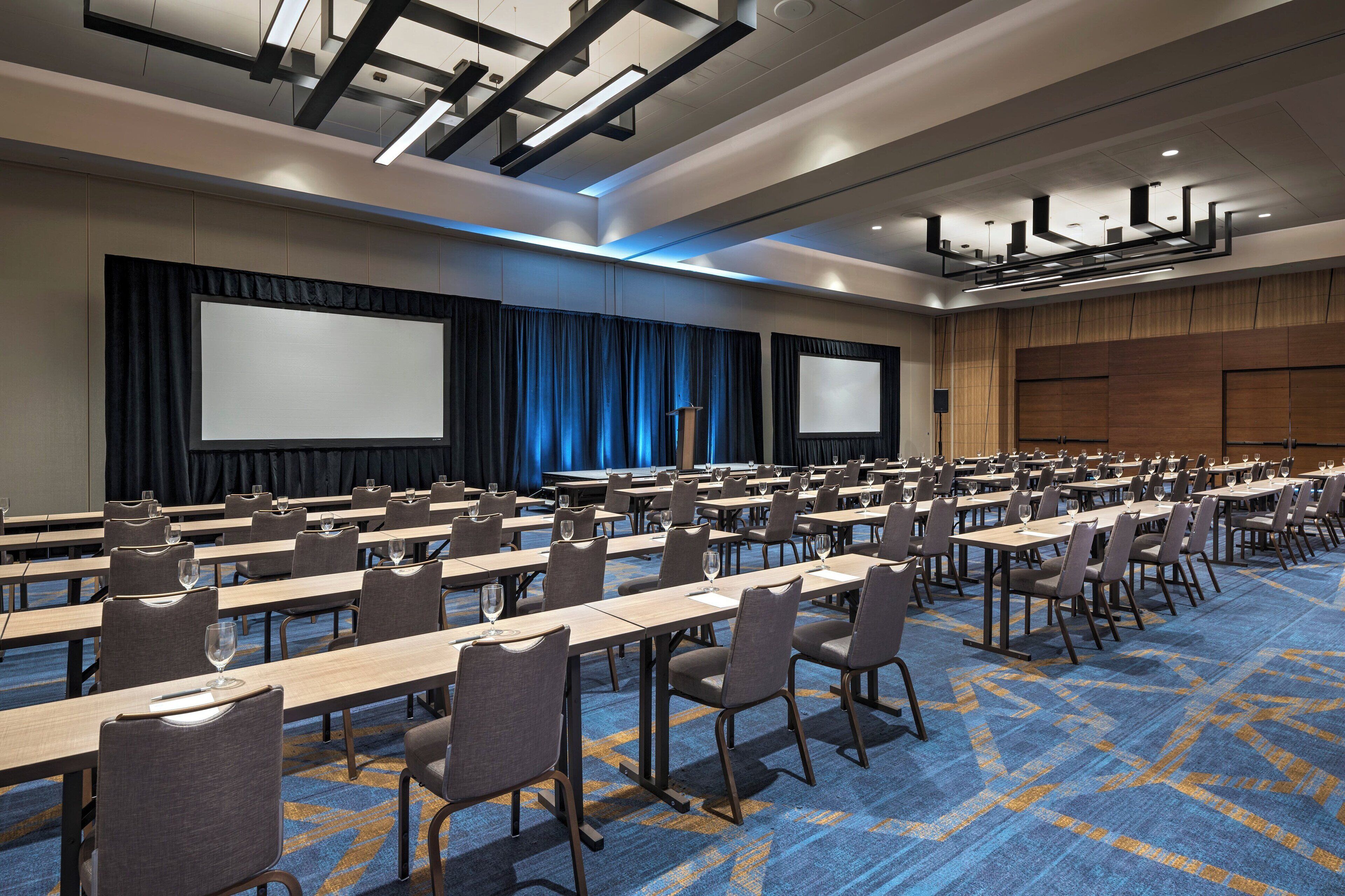 A conference room with rows of tables, chairs, and screens, equipped with projectors and a podium.