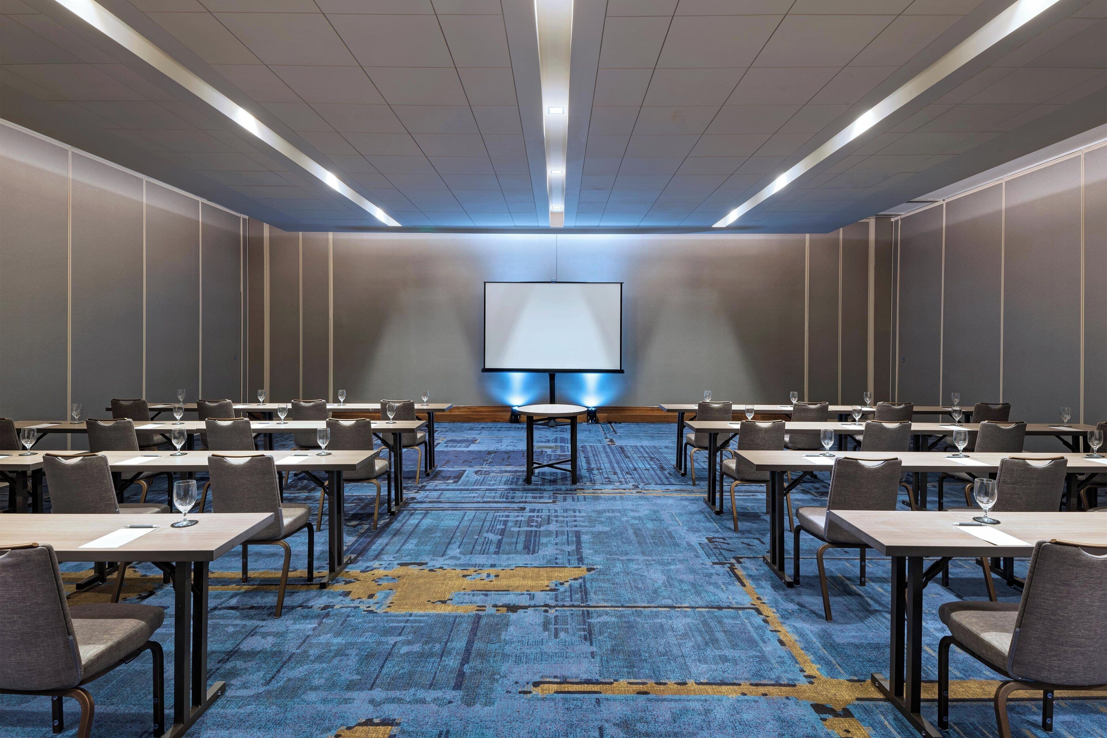 Un salón de conferencias con una pantalla grande, varias mesas y sillas dispuestas para una reunión.
