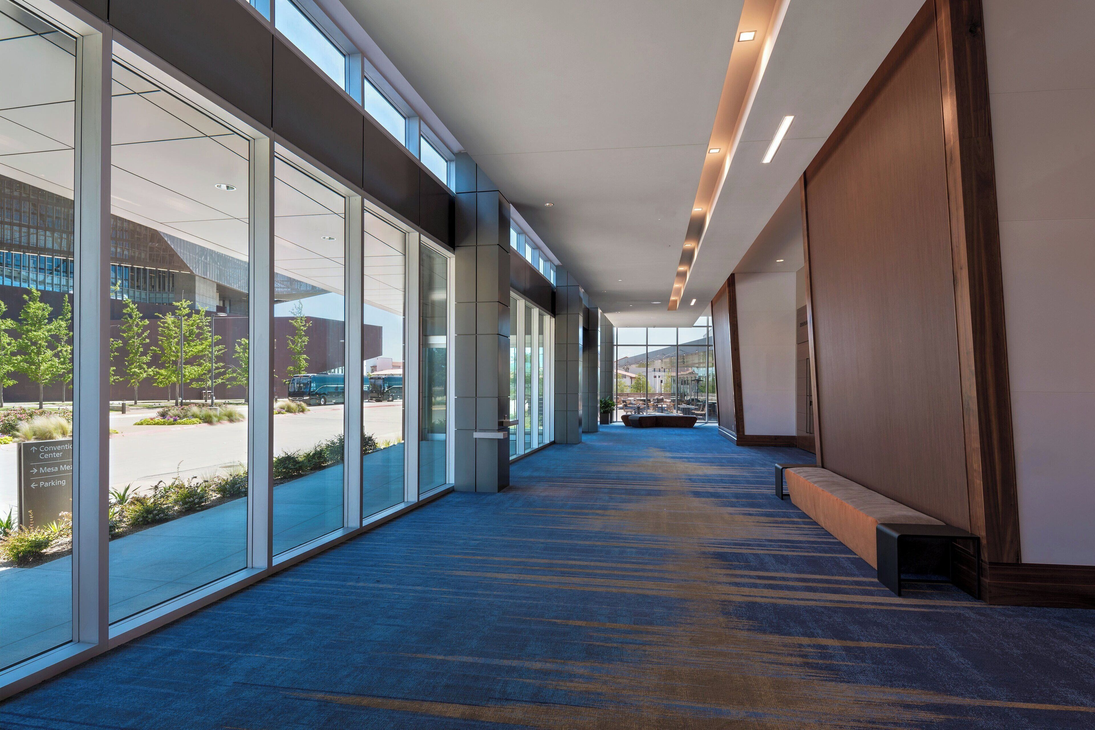 A modern hotel corridor with glass walls, a blue carpet, and a wooden bench.