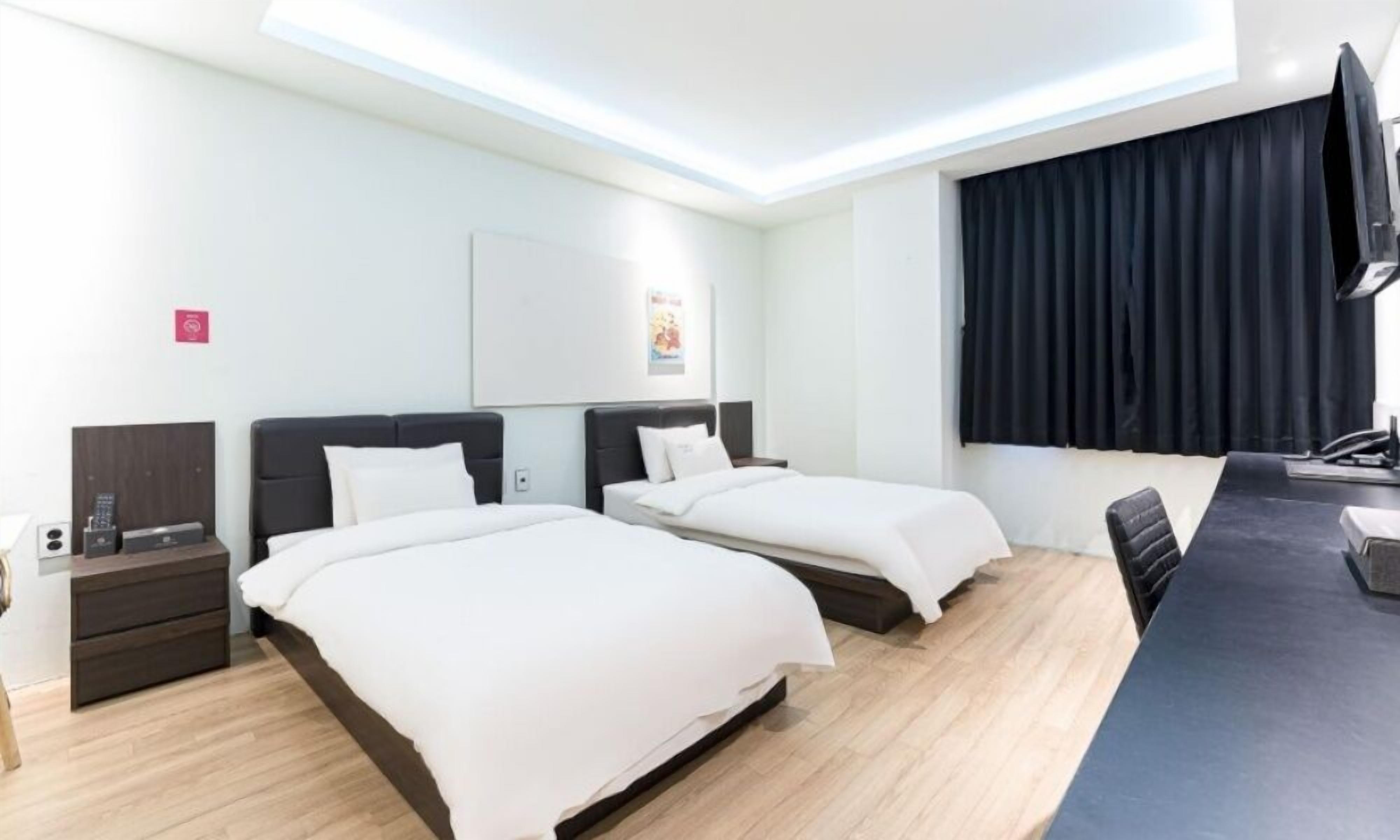 A hotel room with two beds, a desk, a chair, a flat-screen TV, and a window with curtains.