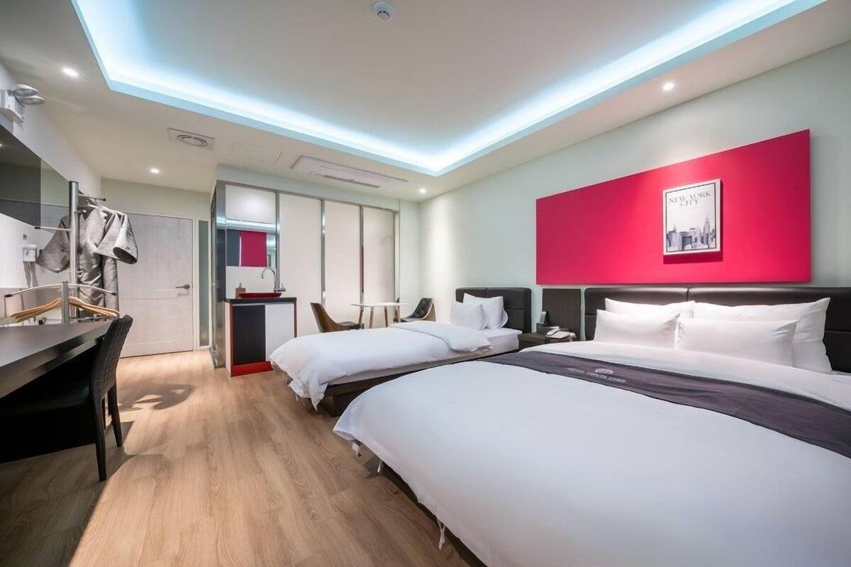A modern hotel room with two beds, a desk, a chair, and a red accent wall.