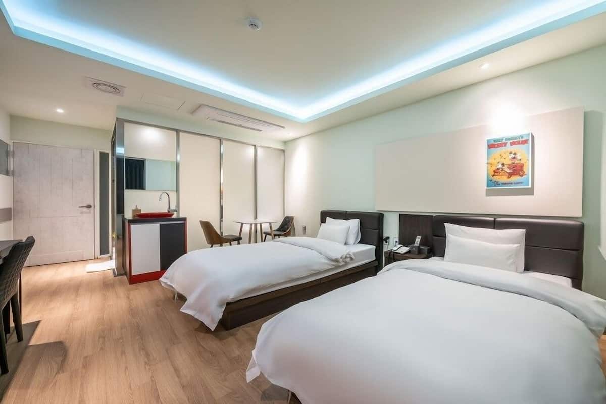 A hotel room with two beds, a desk with a red sink, a chair, and a poster on the wall.