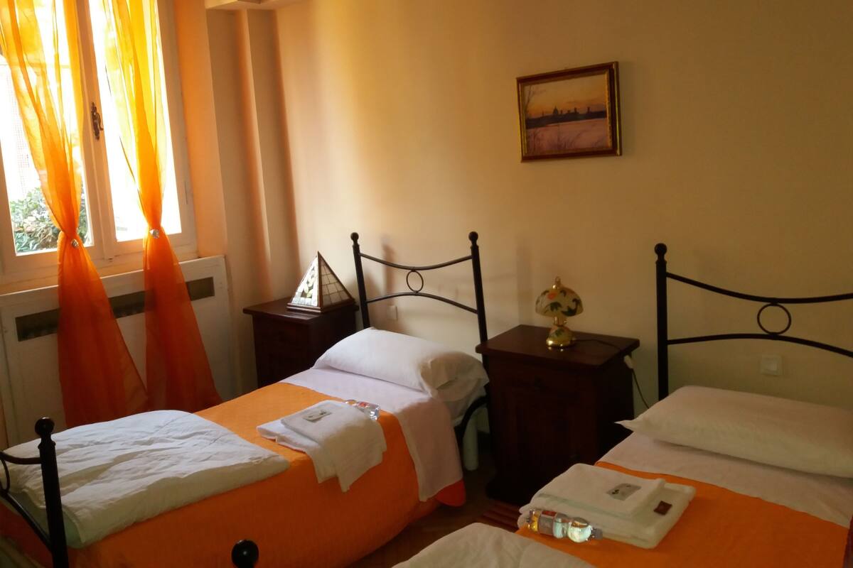 A hotel room with two beds, a nightstand with a lamp, a painting on the wall, and a window with curtains.