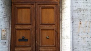 Uma porta dupla de madeira com puxador de metal e fechadura, emoldurada em uma estrutura de concreto com parede pintada de branco e uma placa com o número 27.